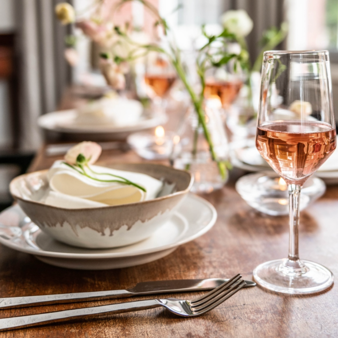 A close-up of an elegant table setting featuring Simon Pearce Burlington Dinnerware Bluff, a bowl, napkin, fork, knife, and a glass of rosé on a wooden table with flowers in soft natural light for an organic look.