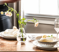 A wooden table set with Simon Pearce Burlington Dinnerware Bluff, cloth napkins, glassware, a clear vase of pale flowers, and a pottery bowl with a small flower sits before a sunlit window draped in sheer curtains for an organic look.