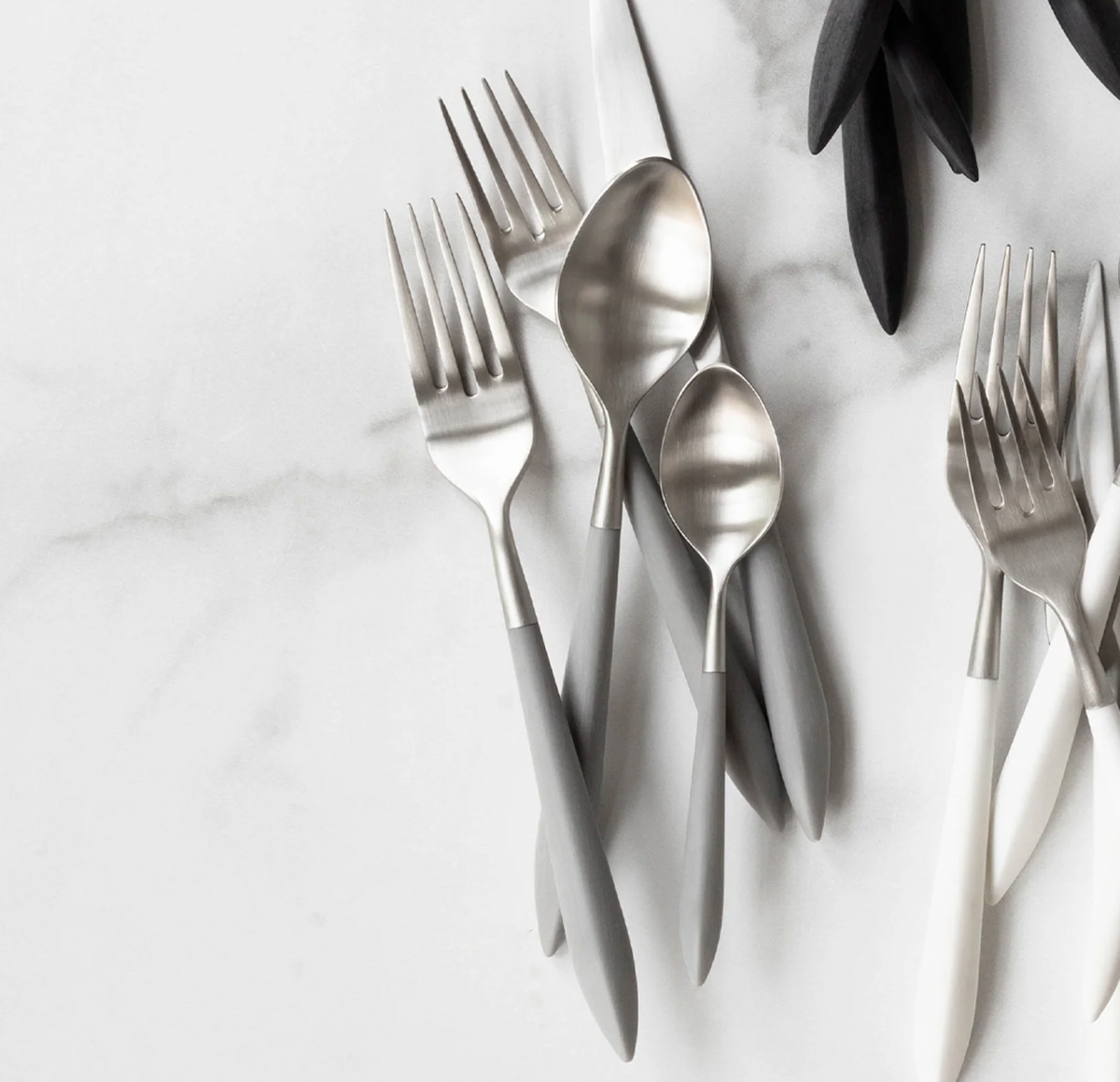 The Vietri Ares Oro Flatware 5 Piece Setting - Grey features matte silver and gray forks, spoons, and a knife with stainless steel construction; some black-handled utensils also appear in the top right on a white marble surface.