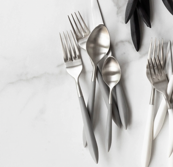 The Vietri Ares Oro Flatware 5 Piece Setting - Grey features matte silver and gray forks, spoons, and a knife with stainless steel construction; some black-handled utensils also appear in the top right on a white marble surface.