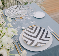 A table set for a formal meal with bold black and white geometric plates, six wine glasses, Vista Alegre Domo Matt Gold Flatware 5 Piece Setting, and a bouquet of white flowers on a light blue tablecloth.