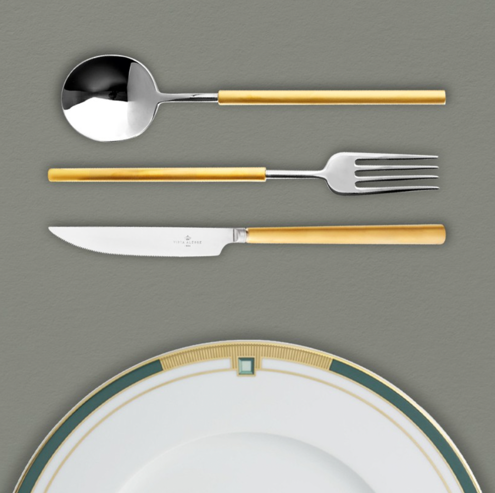 The Domo Matt Gold Flatware 5 Piece Setting by Vista Alegre, with yellow handles, is arranged above a plate with a green, gold, and white border on a gray surface.