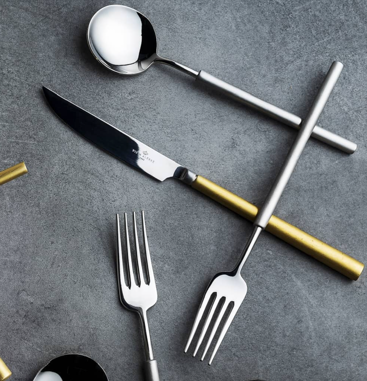The Vista Alegre Domo Matt Gold Flatware 5 Piece Setting, featuring metallic and gold handles, is elegantly arranged on a dark gray textured surface.