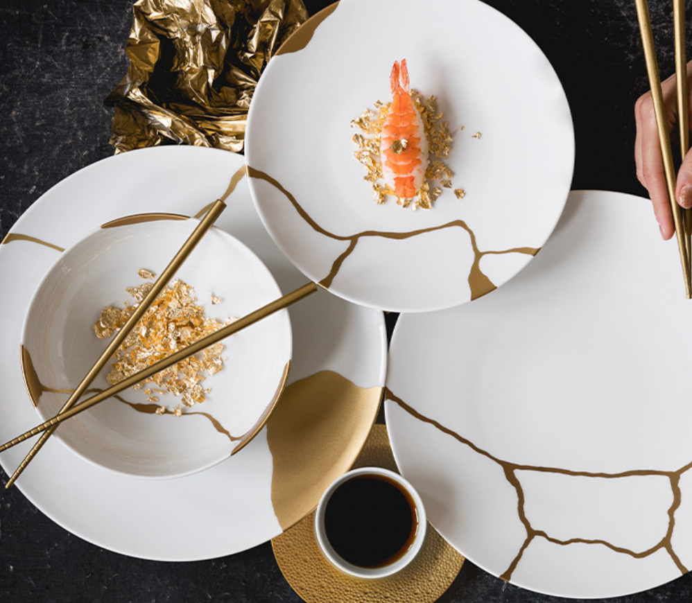 A hand uses gold chopsticks to pick up sushi from a Bernardaud Kintsugi Dinnerware plate, featuring gold cracks, with matching gold-accented dishes, edible gold flakes, and soy sauce on a black table.