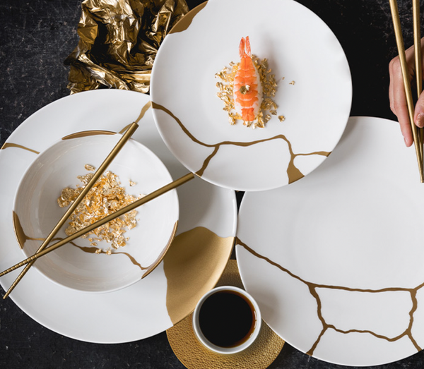 A hand uses gold chopsticks to pick up sushi from a Bernardaud Kintsugi Dinnerware plate, featuring gold cracks, with matching gold-accented dishes, edible gold flakes, and soy sauce on a black table.