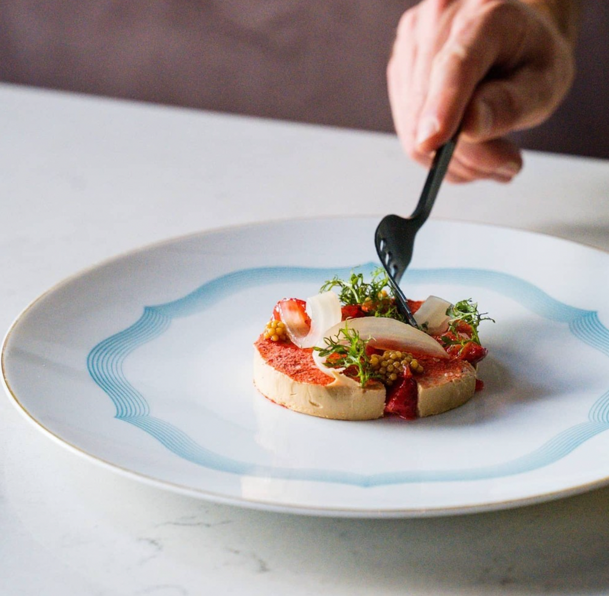 A hand with a black fork is about to enjoy a gourmet dish served on Raynaud's Aura Dinnerware, featuring layered ingredients and greens artfully presented on the scalloped white plate atop a marble surface.
