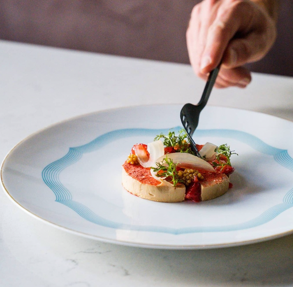 A hand with a black fork is about to enjoy a gourmet dish served on Raynaud's Aura Dinnerware, featuring layered ingredients and greens artfully presented on the scalloped white plate atop a marble surface.