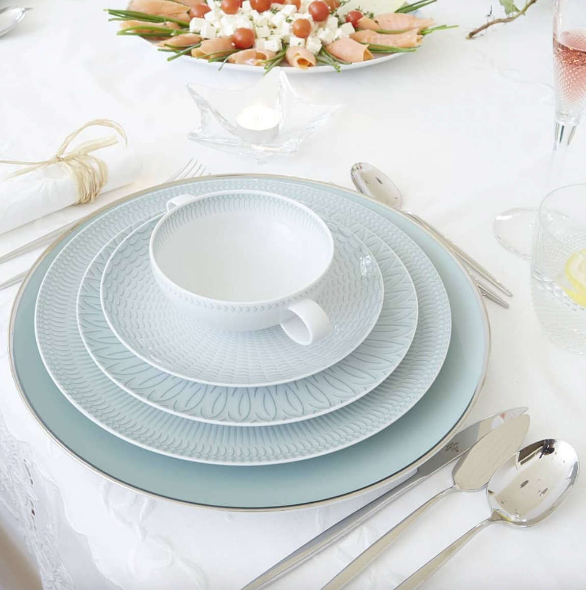 A formal table is set with Vista Alegre Trasso Dinnerware, silver cutlery, a teacup, pink drink, lemon water, and a platter of rolled vegetable appetizers atop a white tablecloth.