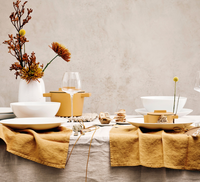 A table set for a meal features Tac 02 White Dinnerware by Jars - Rosenthal, wine glasses, golden-yellow napkins, a beige tablecloth, and a white vase with yellow and orange flowers, all against a neutral background.