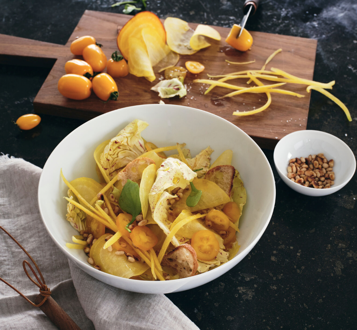 A salad of yellow tomatoes, beet slices, cabbage, and pine nuts is served in a Tac 02 White Dinnerware bowl by Jars - Rosenthal, on a dark counter next to a small bowl of pine nuts and a wooden board with sliced yellow tomatoes and a peeler.