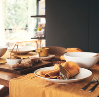 A wooden dining table set features Jars - Rosenthal Tac 02 White Dinnerware, yellow-orange linens, napkins, cutlery, a bread loaf on a board, and bowls. Warm natural light from the window creates a cozy, inviting atmosphere.