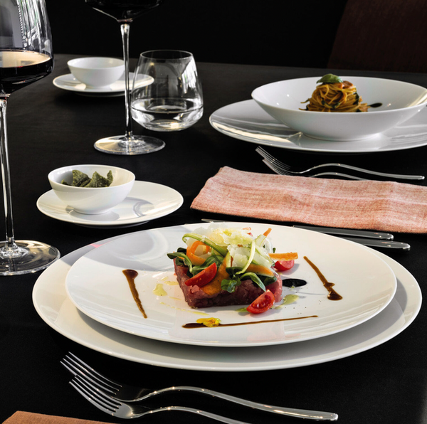 A gourmet meal is artfully plated on Tac 02 White Dinnerware from Jars - Rosenthal, with diced tuna, cherry tomatoes, greens, and sauce. A glass of red wine, pasta bowl, and napkin are arranged on a black tablecloth.