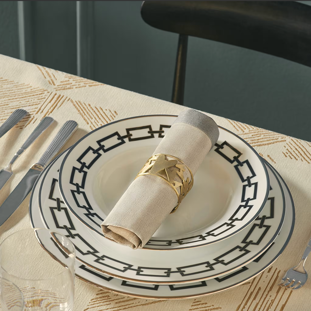 A table setting with two Ginori 1735 Catene Zaffiro porcelain plates, bold black borders, a beige napkin in a gold geometric ring, silver cutlery, a glass, and a patterned tablecloth.
