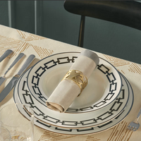A table setting with two Ginori 1735 Catene Zaffiro porcelain plates, bold black borders, a beige napkin in a gold geometric ring, silver cutlery, a glass, and a patterned tablecloth.