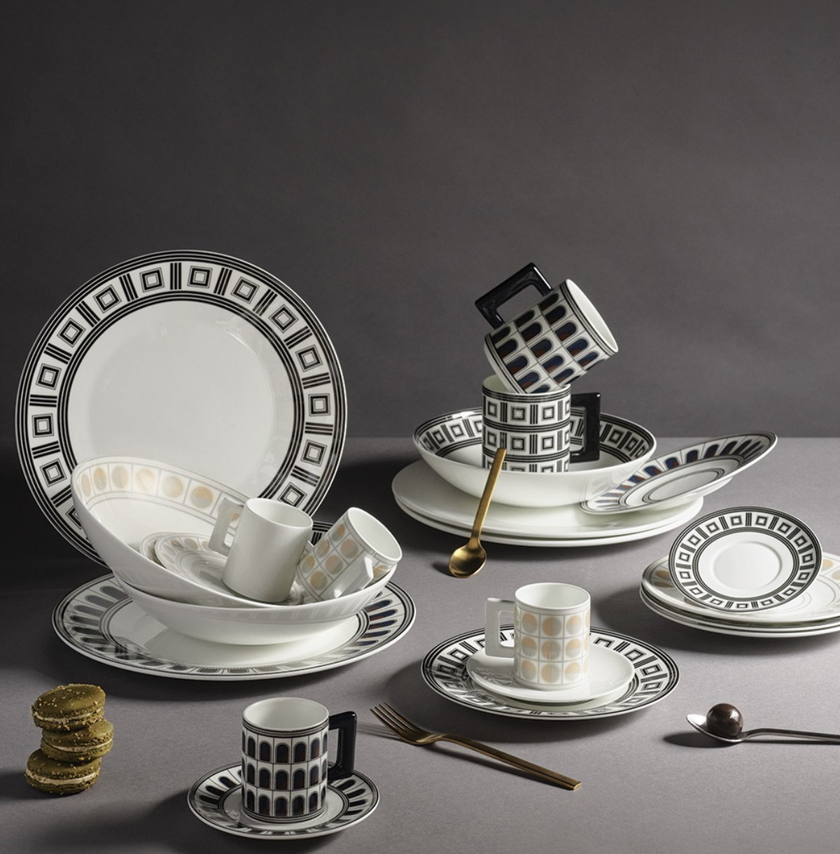 A Greg Natale Palazzo Navy Dinnerware 3 Piece Set of plates, bowls, and cups—some stacked—crafted from fine bone china sits with a gold spoon, fork, pastel macarons, and a silver spoon on a grey background.