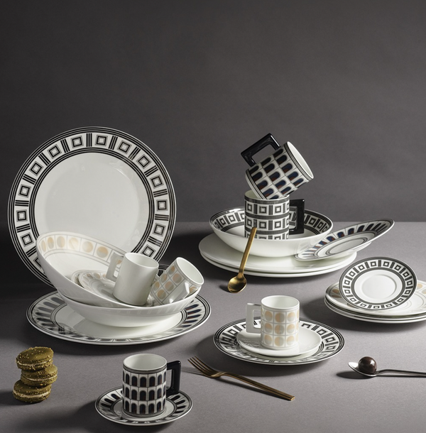 A Greg Natale Palazzo Navy Dinnerware 3 Piece Set of plates, bowls, and cups—some stacked—crafted from fine bone china sits with a gold spoon, fork, pastel macarons, and a silver spoon on a grey background.