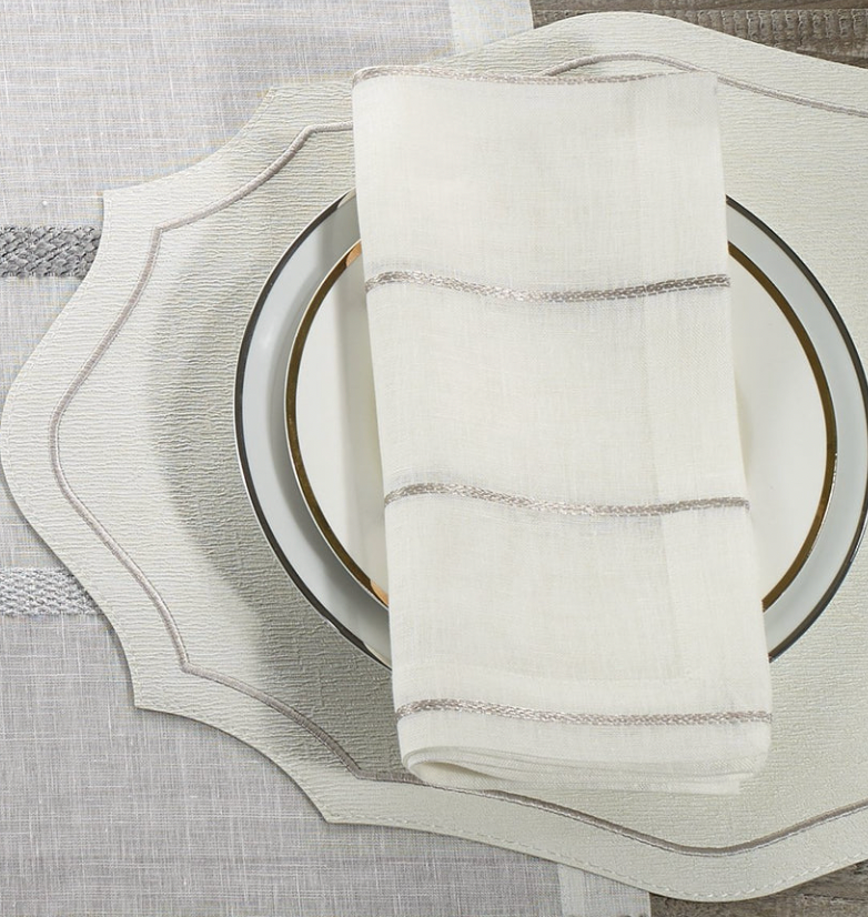 The Bodrum Metallic Thread Napkin Silver Set of 4 features white and silver-striped soft linen napkins presented on a gold-rimmed plate atop a white and beige scalloped placemat over a textured neutral tablecloth.