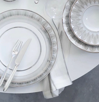 Elegant white and silver patterned plates, a fork and knife on the Bodrum Metallic Thread Napkin Silver Set of 4, with glassware arranged neatly on a round white table atop a gray floor.