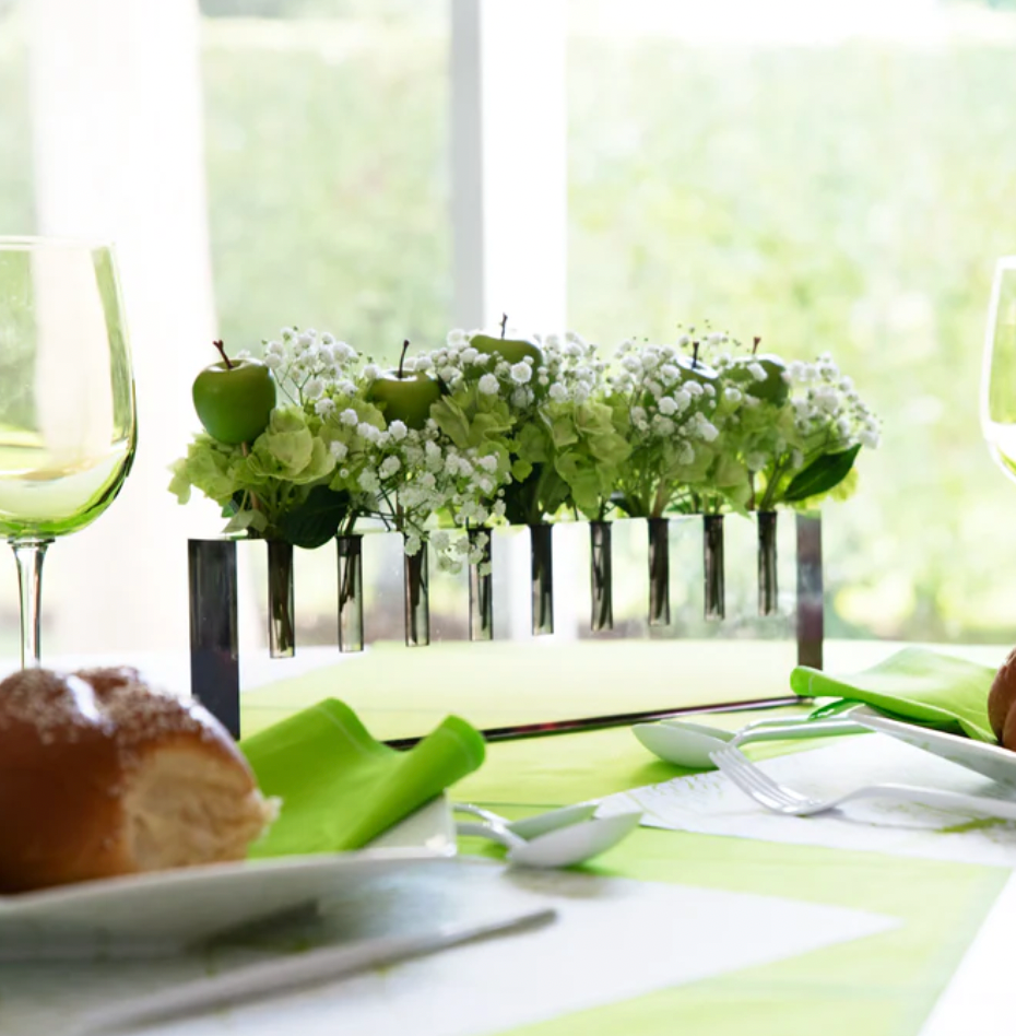 A table setting featuring the Apeloig Modern Acrylic Oil Menorah as a centerpiece, complemented by a bread roll and a green wine glass.