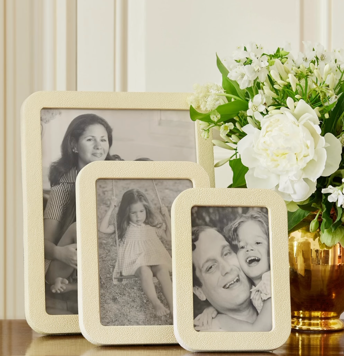 Three family photos in Aerin Modern Shagreen Frame Creams sit next to a gold vase with white flowers and greenery, arranged on a surface against a light paneled wall.