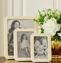Three family photos in Aerin Modern Shagreen Frame Creams sit next to a gold vase with white flowers and greenery, arranged on a surface against a light paneled wall.