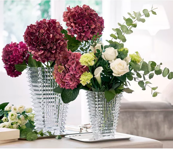 A vibrant floral arrangement—deep red and pink hydrangeas, white roses, green blooms, and eucalyptus—adorns a bright living room in the striking Baccarat Eye Rectangular Vase Clear.