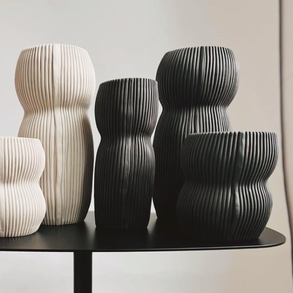 The Curvy Round Vase Black by CYM Warkov Ceramics, with its vertical grooves and organic curves, is arranged on a dark tabletop against a neutral background, highlighting the elegance of artisan pottery.