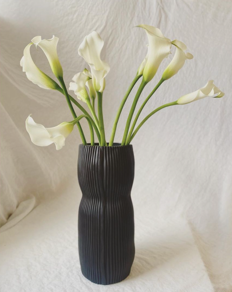 The Curvy Round Vase Black by CYM Warkov Ceramics holds long-stemmed white calla lilies, set against a soft, light-colored fabric background.