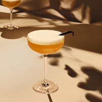A Richard Brendon Fluted Coupe holding a frothy yellow cocktail with a vanilla bean garnish sits on a beige surface, casting artistic shadows; another Fluted Coupe is softly blurred in the background.