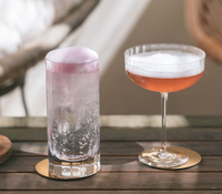 A purple bubbly drink in a Fluted Coupe by Richard Brendon and a pink foamy cocktail sit elegantly on round gold coasters atop a wooden table.