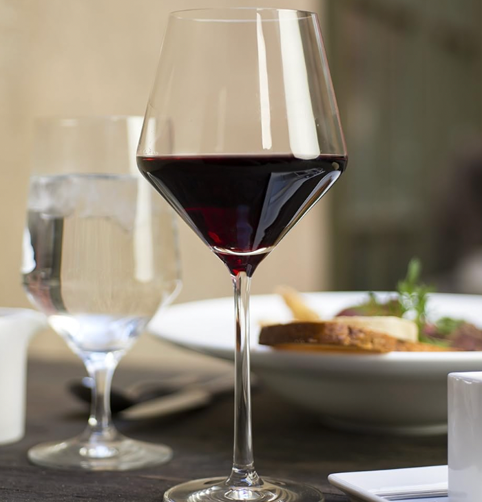 A glass of red wine in Zwiesel Fortessa’s Zweisel Pure Barware sits next to a glass of water and a white bowl of food, with a blurred dining setting in the background.