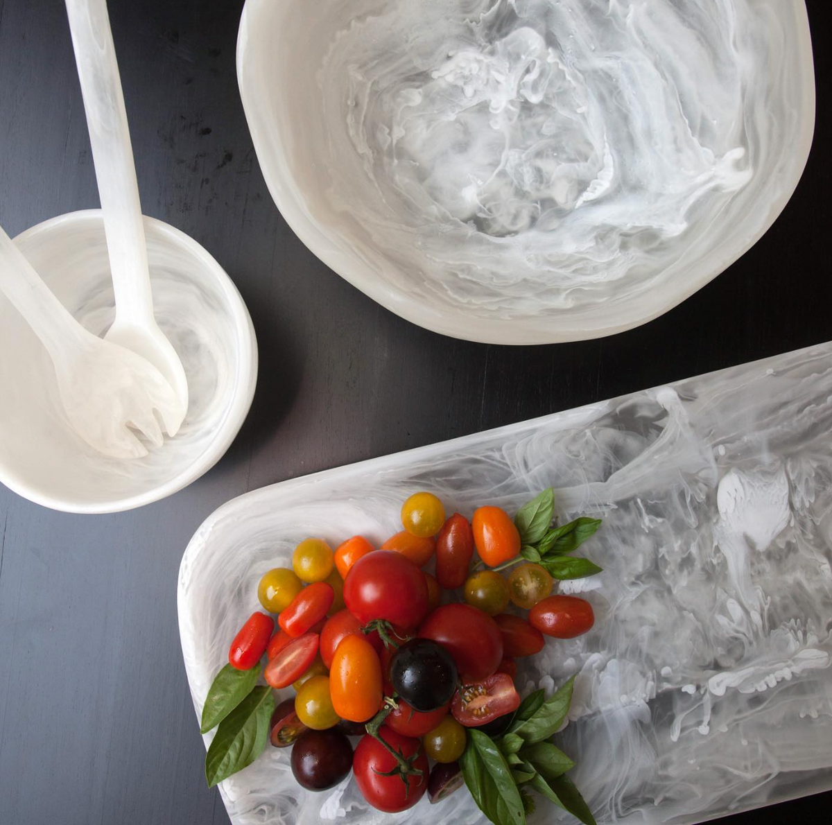 Colorful cherry tomatoes and fresh basil rest on the Nashi Home Swirl Classic Resin Rectangle Platter, with empty white marbled bowls and utensils arranged around them on a dark tabletop.