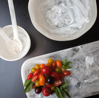 Colorful cherry tomatoes and fresh basil rest on the Nashi Home Swirl Classic Resin Rectangle Platter, with empty white marbled bowls and utensils arranged around them on a dark tabletop.