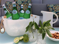 A white, dishwasher-safe ice bucket holds sparkling water next to the Nashi Home Swirl Resin Classic Pitcher, glasses with mint and lime, and a bowl of nuts. Patterned pillows accent the sofa in the background.