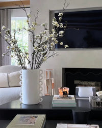 A modern living room with a black coffee table featuring the Tina Frey Loopy Resin Vase White filled with flowering branches, stacked books, a lit candle, and a TV mounted above the fireplace in the background.