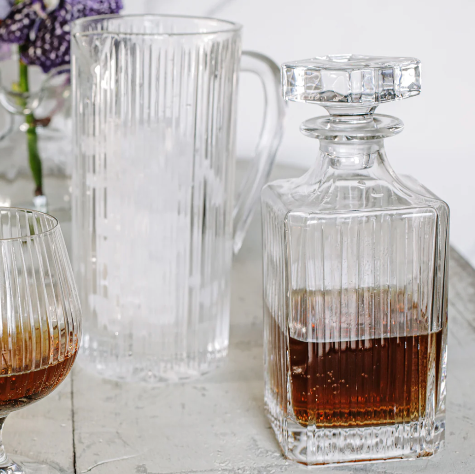 The Vietri Natalia Decanter, filled with whiskey, sits beside an empty pitcher and a half-full snifter on a light table, purple flowers in the background, creating an inviting bar setting.