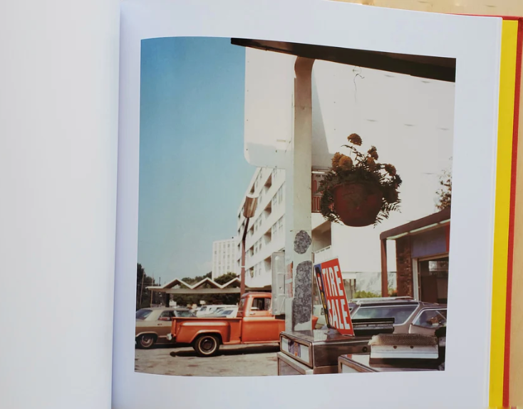A vintage photo in "William Eggleston: 2 1/4" by ARTBOOK DAP/INGRAM captures an orange pickup, silver car, FIRE SALE sign, and hanging flower pot at a gas station—classic color photography beneath a clear blue sky.