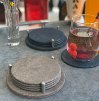 Close-up of Bodrum's Stingray Coaster Set of 4 featuring textured metallic finishes in silver and dark gray, with a glass of fruit water on one, all atop a gray table. Easy-care design makes them ideal for daily use.