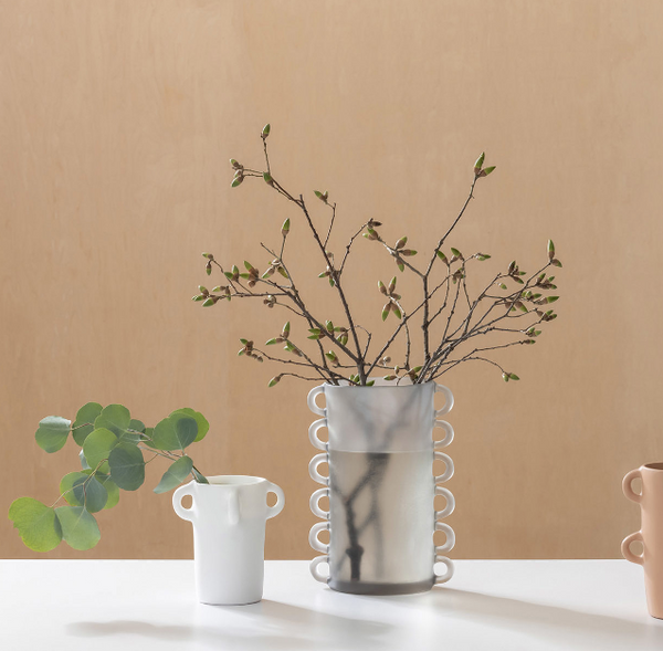 A minimalist arrangement featuring the tall handmade Loopy Vase Fog by Tina Frey with budding branches, a small white cup with round handles and eucalyptus leaves, and a partial peach mug, all on a white surface against a beige background.
