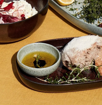 A plate from the L'Objet Terra Seafoam Dinnerware holds a bowl of olive oil and balsamic vinegar, rustic bread, and rosemary on a yellow tablecloth. A salad bowl with fresh herbs is partially visible in the background.