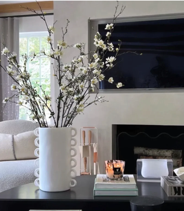 A modern living room features a black coffee table with the Tina Frey Loopy Vase Fog holding flowering branches, a lit candle, and stacked books. Behind it are a fireplace, TV, and a light-colored sofa.