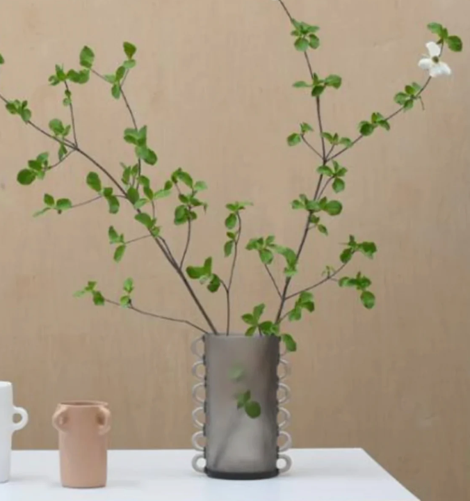 A minimalist arrangement features leafy branches and a white flower in the Tina Frey Loopy Vase Fog, set on a white surface beside two small ceramic vases, with a plain beige background.