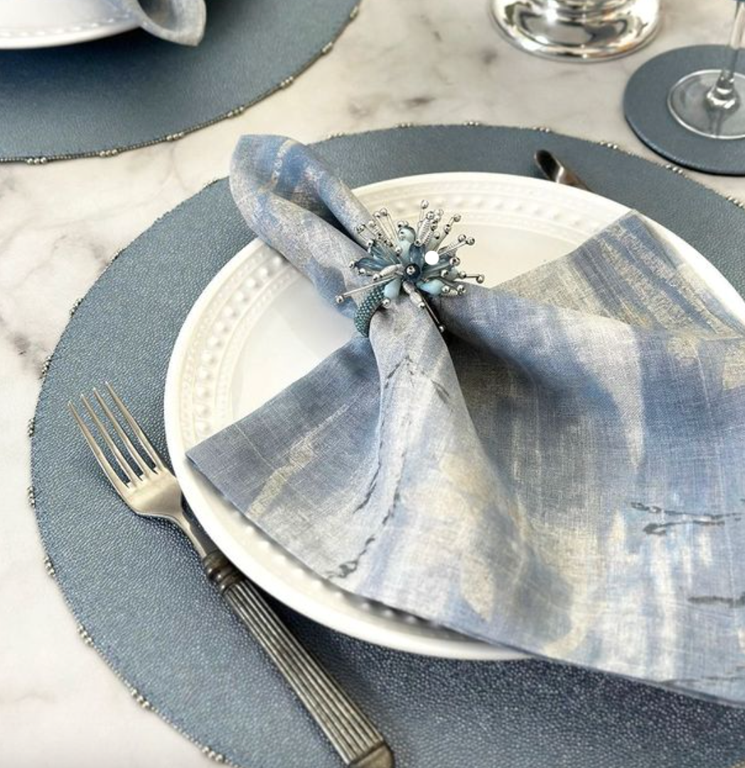 A white plate with a shimmery blue napkin in a beaded ring rests on the Bodrum Charm Placemat Ice Blue. A fork and knife are on the left, with part of a wine glass visible on the right. Set includes 4 placemats.