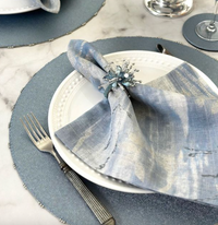 A white plate with a shimmery blue napkin in a beaded ring rests on the Bodrum Charm Placemat Ice Blue. A fork and knife are on the left, with part of a wine glass visible on the right. Set includes 4 placemats.