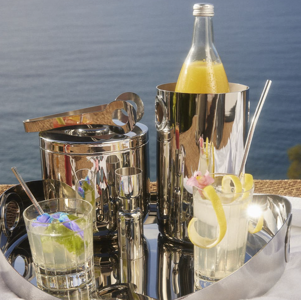 A silver tray displays two cocktails with garnish, the Christofle Oh De Stainless Steel Wine Cooler holding orange juice, a shaker, an ice bucket with tongs, and the ocean beyond—perfect for your home bar.