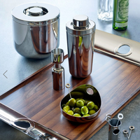 A modern bar setup features the Christofle Oh De Stainless Large Steel Bowl filled with green olives and picks, alongside a wooden tray, cocktail shaker, jigger, matching ice bucket, with a bottle and glasses in the background.