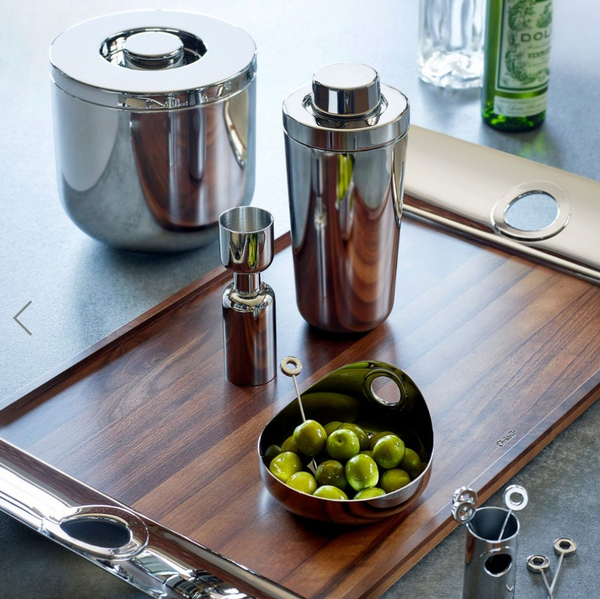 A modern bar setup features the Christofle Oh De Stainless Large Steel Bowl filled with green olives and picks, alongside a wooden tray, cocktail shaker, jigger, matching ice bucket, with a bottle and glasses in the background.