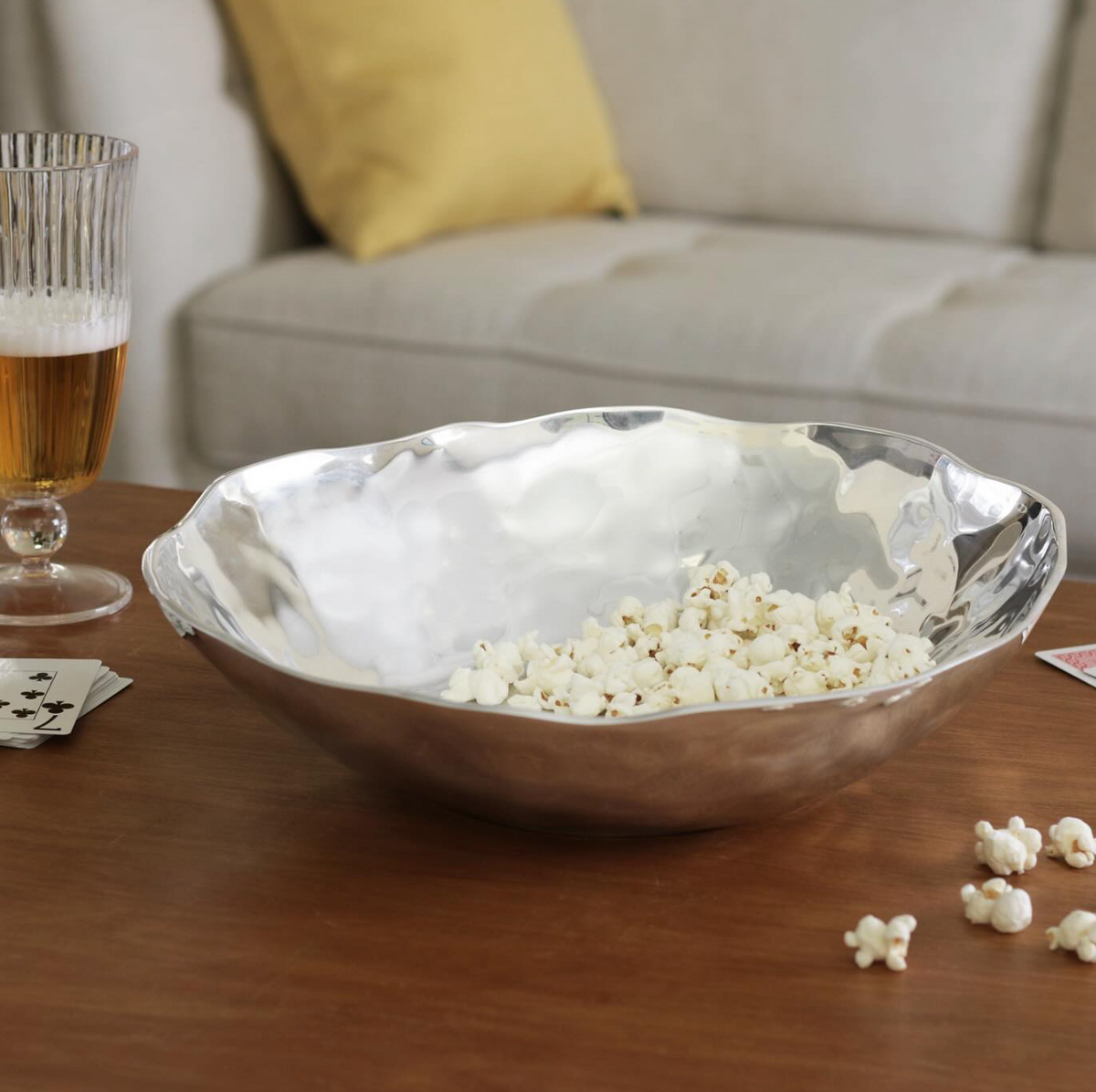 The Beatriz Ball Soho Nueve Organic Bowl 12.5" brims with popcorn atop a wooden coffee table. Scattered playing cards, popcorn pieces, a glass of beer, and a beige couch with yellow pillows complete the inviting scene.