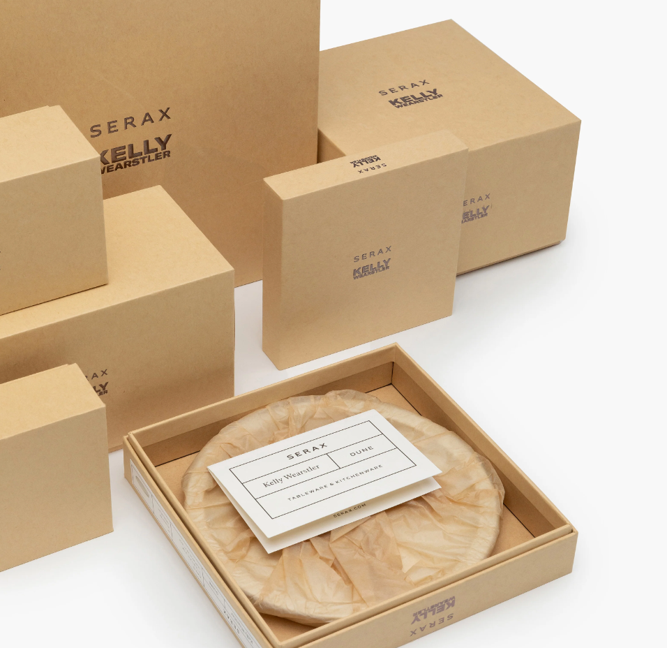 Several tan cardboard boxes labeled Serax are neatly stacked. One open box reveals Zuma Dinnerware Pacific—designer porcelain plates—inspired by Bauhaus style, with an informational card placed on top.