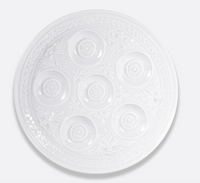 The Bernardaud Louvre Condiment Tray is a round, white, ornate plate with seven circular indentations and intricate patterns, inspired by classic Limoges France craftsmanship.